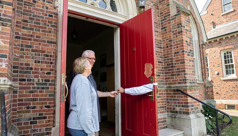 A couple in casual clothes welcomed at the open red front doors of a brick church by an older usher with a warm smile.