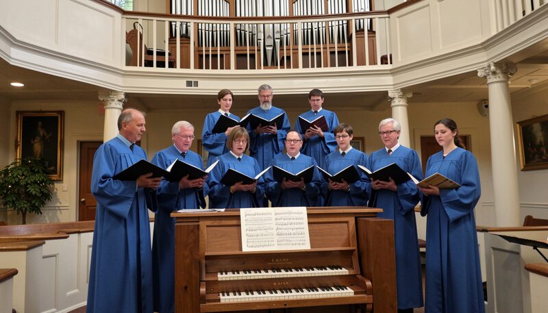 A volunteer choir of eight singers in blue robes holding hymnals in two rows beside a small pipe organ.