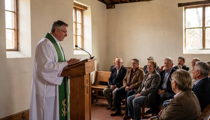 A pastor in a white alb and green stole standing at a wooden lectern reading to a small seated congregation in a simple sanctuary.